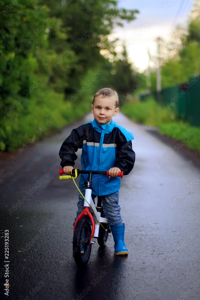 The little boy in a blue raincoat riding a bike on wet asphalt in early autumn The little boy in a blue raincoat riding a bike on wet asphalt in early autumn