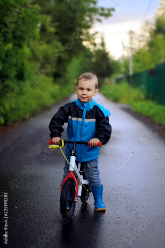 The little boy in a blue raincoat riding a bike on wet asphalt in early autumn
