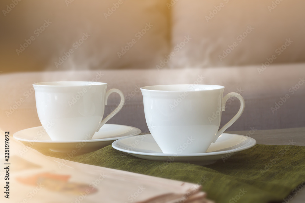 Two white coffee cups with soft smoke stream put on plate and dark green cloth nearly newspaper in the living room and sun lighting from window. Selective focus. Two white coffee cups with soft smoke stream put on plate and dark green cloth nearly newspaper in the living room and sun lighting from window. Selective focus.