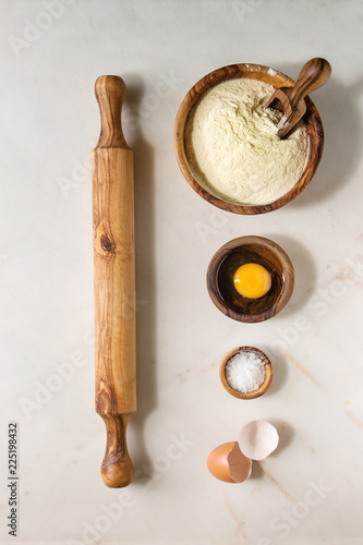 Ingredients for homemade italian pasta ravioli cooking semolina flour, egg yolk, sea salt with olive wood utensils bowls, scoop and rolling pin over white marble background. Flat lay, space