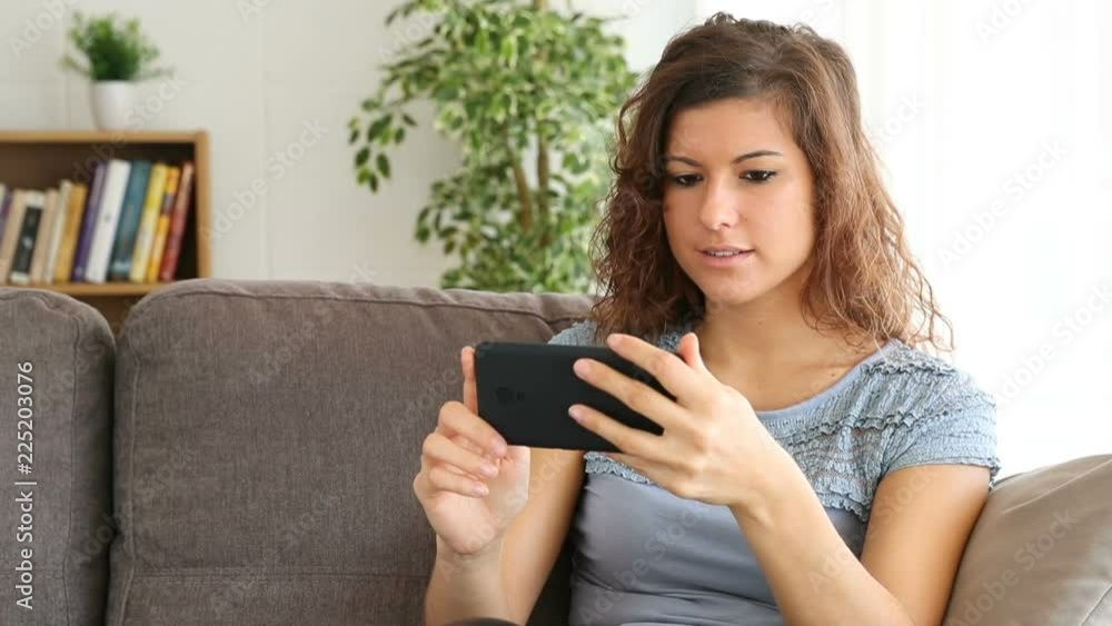 Happy girl watching media content in a smart phone after choosing it sitting on a couch at home Happy girl watching media content in a smart phone after choosing it sitting on a couch at home