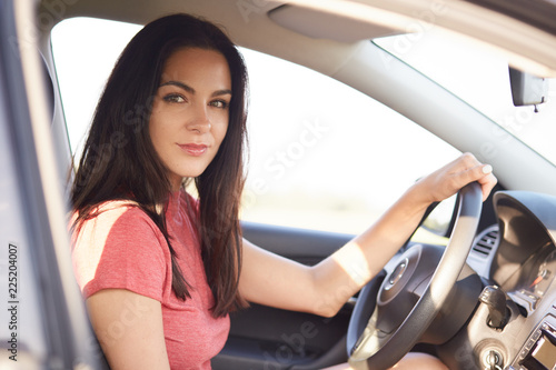 Sideways shot of good looking serious brunette girl drives car professionaly, sits on drivers seat, keeps hand on wheel, ready to cover long distance, dressed in casual clothing. Riding concept © sementsova321