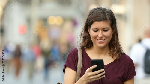 Front view of a happy woman walking towards camera and using a smart phone in the street