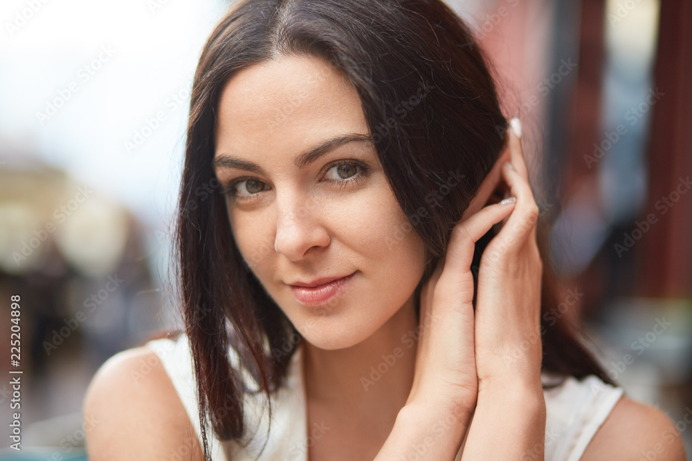 Headshot of pretty woman with dark hair, pleasant appearance, looks directly at camera, poses outdoor, expresses positive feelings. Brunette lady keeps hands together, models for fashion magazine Headshot of pretty woman with dark hair, pleasant appearance, looks directly at camera, poses outdoor, expresses positive feelings. Brunette lady keeps hands together, models for fashion magazine