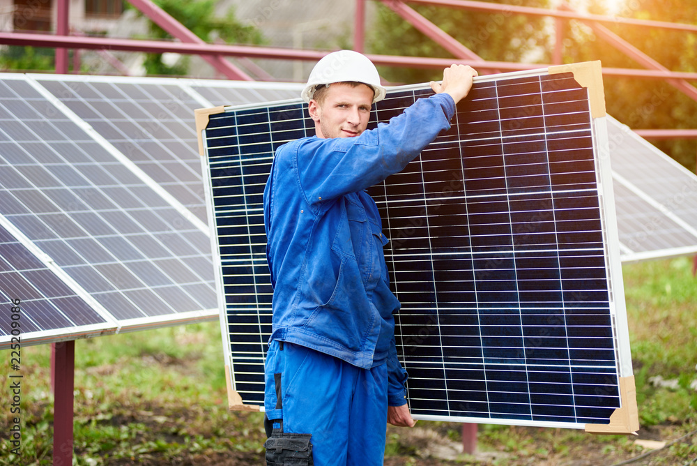 Young worker in blue uniform and protective helmet carrying big shiny solar photo voltaic panel to almost finished exterior metal platform on sunny summer warm day. Renewable ecological green energy. Young worker in blue uniform and protective helmet carrying big shiny solar photo voltaic panel to almost finished exterior metal platform on sunny summer warm day. Renewable ecological green energy.