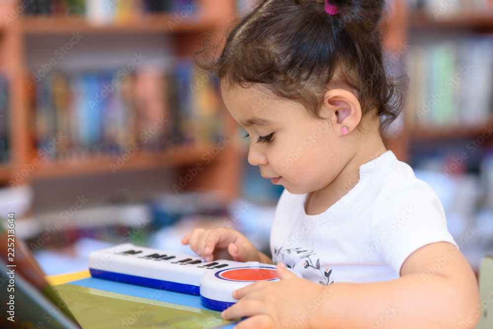 Little Child Indoors In Front Of Books. Cute Young Toddler Sitting On A Chair Near Table and play with toy piano. Kid in a bookstore, surrounded by colorful books. Happy multirace girl play the piano. Little Child Indoors In Front Of Books. Cute Young Toddler Sitting On A Chair Near Table and play with toy piano. Kid in a bookstore, surrounded by colorful books. Happy multirace girl play the piano.