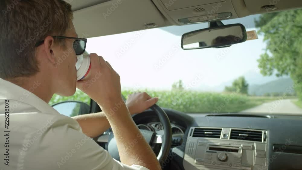 SLOW MOTION, CLOSE UP, COPY SPACE: Young Caucasian man drinks coffee while driving down the empty country road on a beautiful sunny day. Handsome guy on road trip takes a sip of hot tea from paper cup SLOW MOTION, CLOSE UP, COPY SPACE: Young Caucasian man drinks coffee while driving down the empty country road on a beautiful sunny day. Handsome guy on road trip takes a sip of hot tea from paper cup