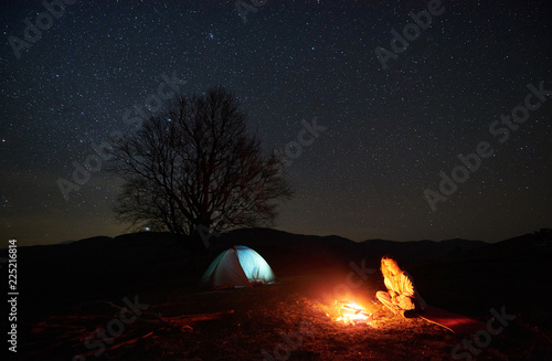 Camping night in mountains. Young female tourist resting near burning bonfire under beautiful starry sky with brightly lit tent and silhouette of big tree in background. Outdoor activity concept