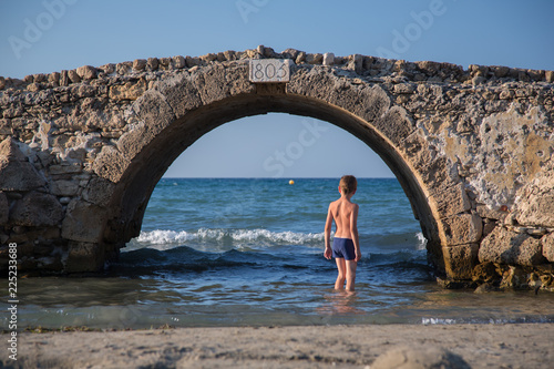 Young boy in swimsuit stands under an old stone bridge in water of sea, in Greece  photo