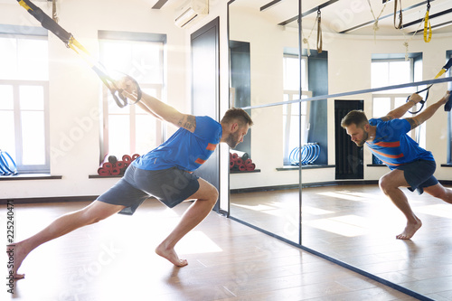 Handsome muscular man athlete doing exercise alone with fitness belts in modern gym club, workout single sport, sun shining.