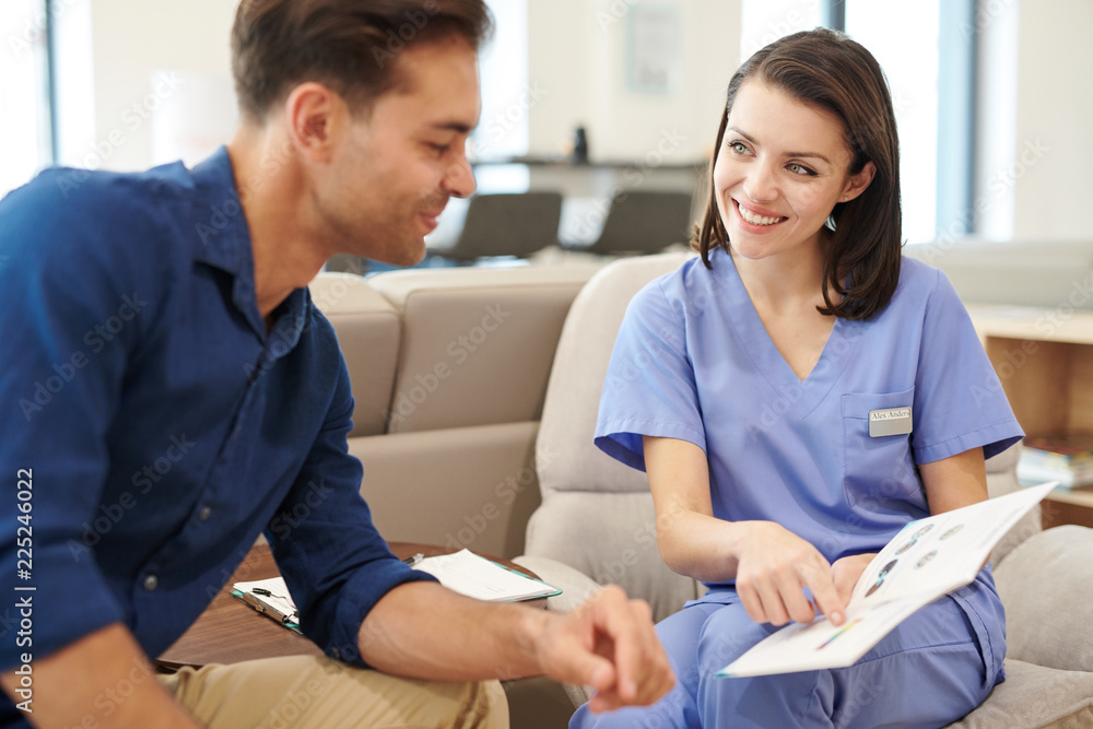 Fototapeta premium Portrait of smiling young nurse offering brochure to patient in hall of modern clinic, copy space
