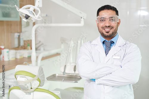 Waist up portrait of confident Middle-Eastern doctor wearing lab coat posing standing with arms crossed in dentists office with dental chair in background, copy space