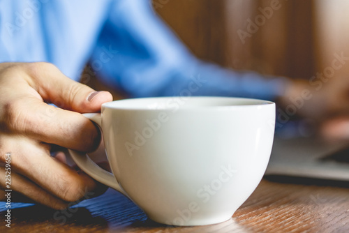 male hand holding a white cup with hot drink on work table whily typing on laptop keyboard f