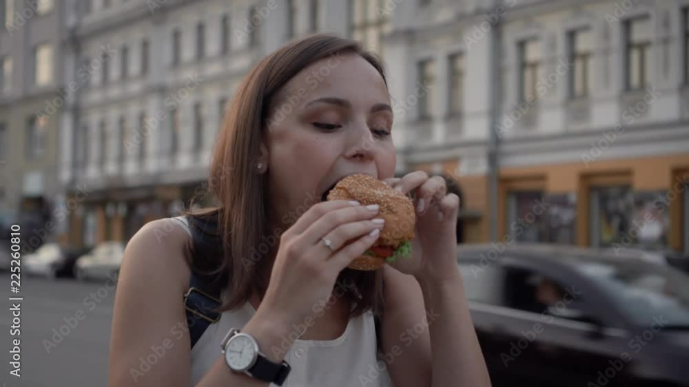 Young attractive woman bites off the hamburger on a city street in the evening. Young attractive woman bites off the hamburger on a city street in the evening.