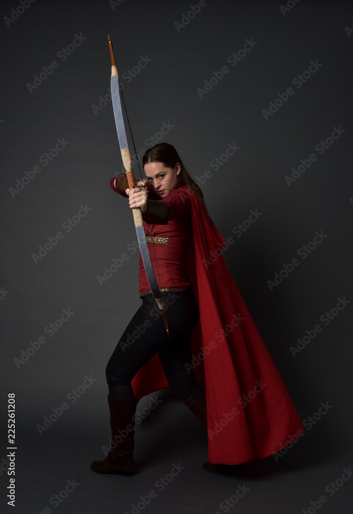 full length portrait of brunette girl wearing red medieval costume and cloak, holding a bow and arrow. standing pose on grey studio background. full length portrait of brunette girl wearing red medieval costume and cloak, holding a bow and arrow. standing pose on grey studio background.