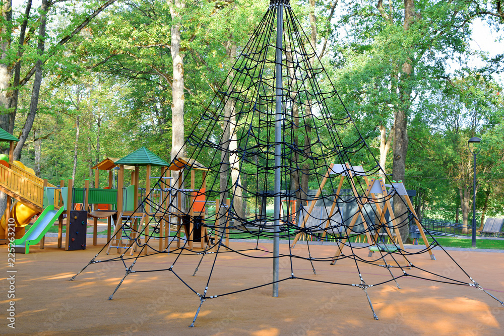 Fototapeta premium Modern playground with climbing net swings and slides with soft protective rubber coating in city park with old trees and pavement pathway in summer day. Children activities outdoors concept.
