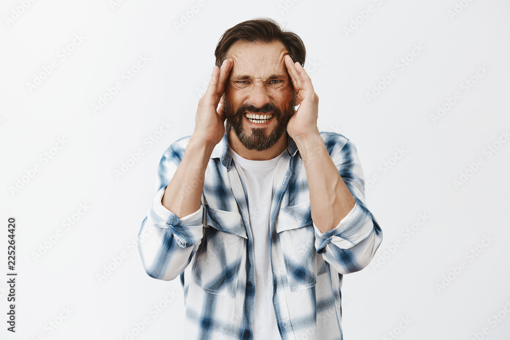 Turn pain off please. Distressed adult bearded guy suffering from painful migraine or headache, grimacing from discomfort, holding hands on temples and staring with unhappy upset look at camera Turn pain off please. Distressed adult bearded guy suffering from painful migraine or headache, grimacing from discomfort, holding hands on temples and staring with unhappy upset look at camera