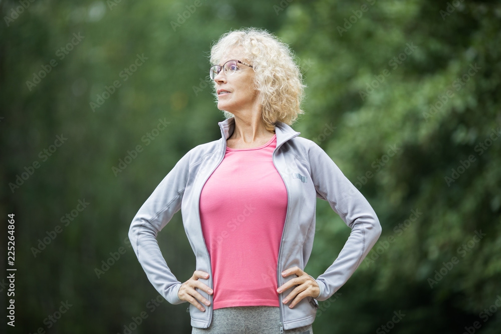 Mature active female in akimbo pose standing in park with green trees behind Mature active female in akimbo pose standing in park with green trees behind
