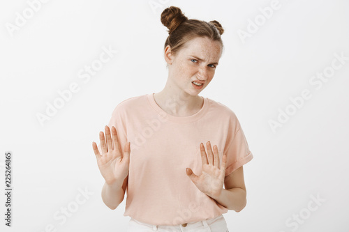 Displeased disgusted female european girlfriend with buns hairstyle and freckles, pulling hands towards camera in rejectiong and no gesture, turning away in dislike, wrinkling nose over grey wall