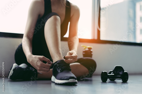 Konstfotografi Wonman sitting shoelaces with protein shake and dumbbell at fitness gym after running exercise workout for cardio and muscle building