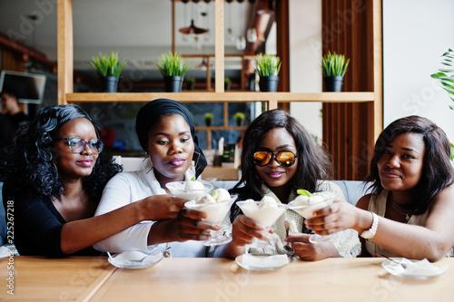 Four african american girls sitting on table at cafe and cheers with ice cream dessert.