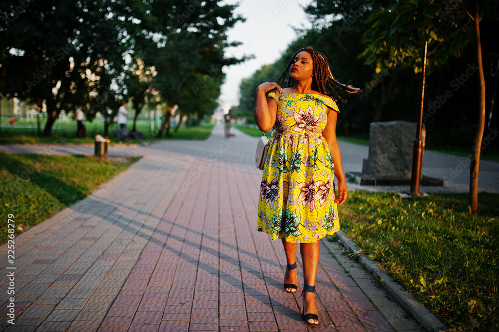 Fototapeta premium Cute small height african american girl with dreadlocks, wear at coloured yellow dress, posed at sunset.