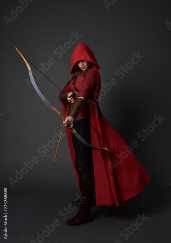 Fotografía full length portrait of brunette girl wearing red medieval costume and cloak, holding a bow and arrow