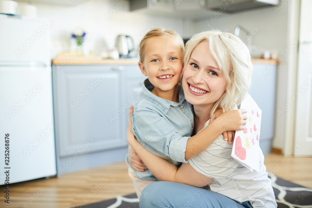 Cheerful little girls and her pretty blonde mother in embrace looking at you in the kitchen Cheerful little girls and her pretty blonde mother in embrace looking at you in the kitchen