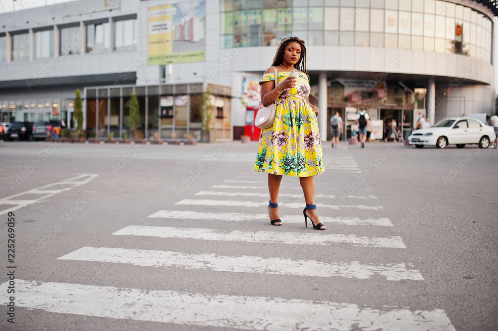Naklejka premium Cute small height african american girl with dreadlocks, wear at coloured yellow dress, walking at crosswalk against trade center with cup of coffee at hand.