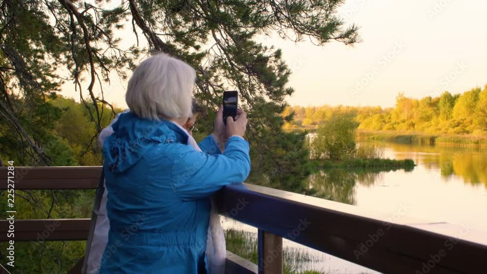 Two senior women photographing river on phone in autumn park Two senior women photographing river on phone in autumn park