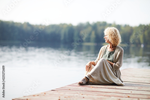 Serious pensive curly-haired mature lady in long skirt sitting on pier and contemplating tranquil nature around