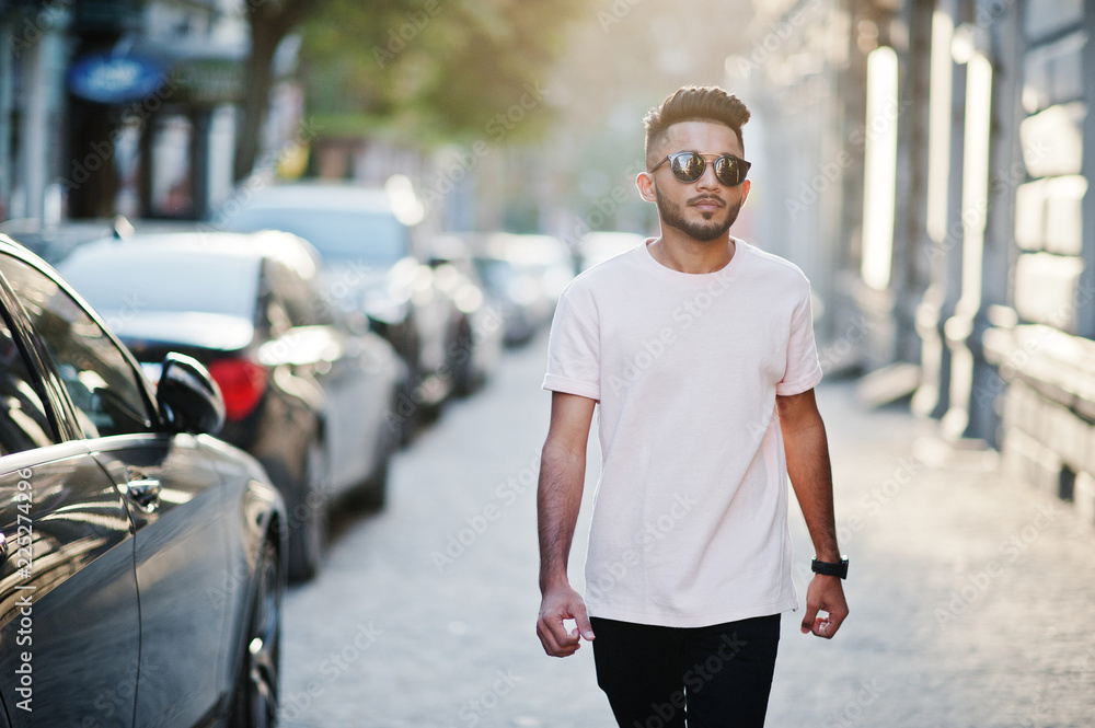 Obraz premium Stylish indian beard man at sunglasses and pink t-shirt. India model walking outdoor sunset at streets of city.