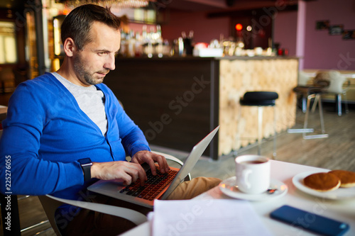 Content enterprising handsome male freelancer in blue cardigan sitting at table and keeping laptop on knees while doing work in cafe during breakfast