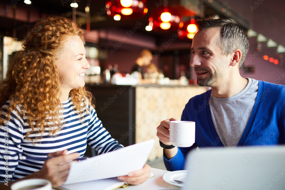 Cheerful successful entrepreneurs sitting at table and discussing papers while planning startup project in cafe Cheerful successful entrepreneurs sitting at table and discussing papers while planning startup project in cafe