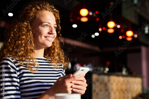 Cheerful excited beautiful girl with curly hair holding smartphone and reading sms from boyfriend while being in anticipation, she resting in cafe