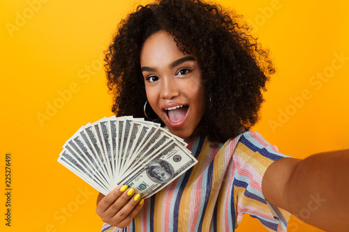 Slika na platnu Excited young african woman posing isolated over yellow background holding money make selfie by camera
