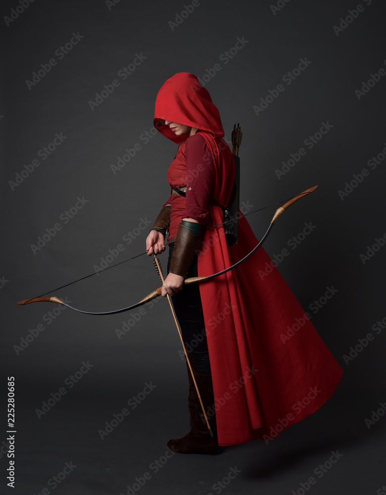 Fototapeta premium full length portrait of brunette girl wearing red medieval costume and cloak, holding a bow and arrow. standing pose on grey studio background.