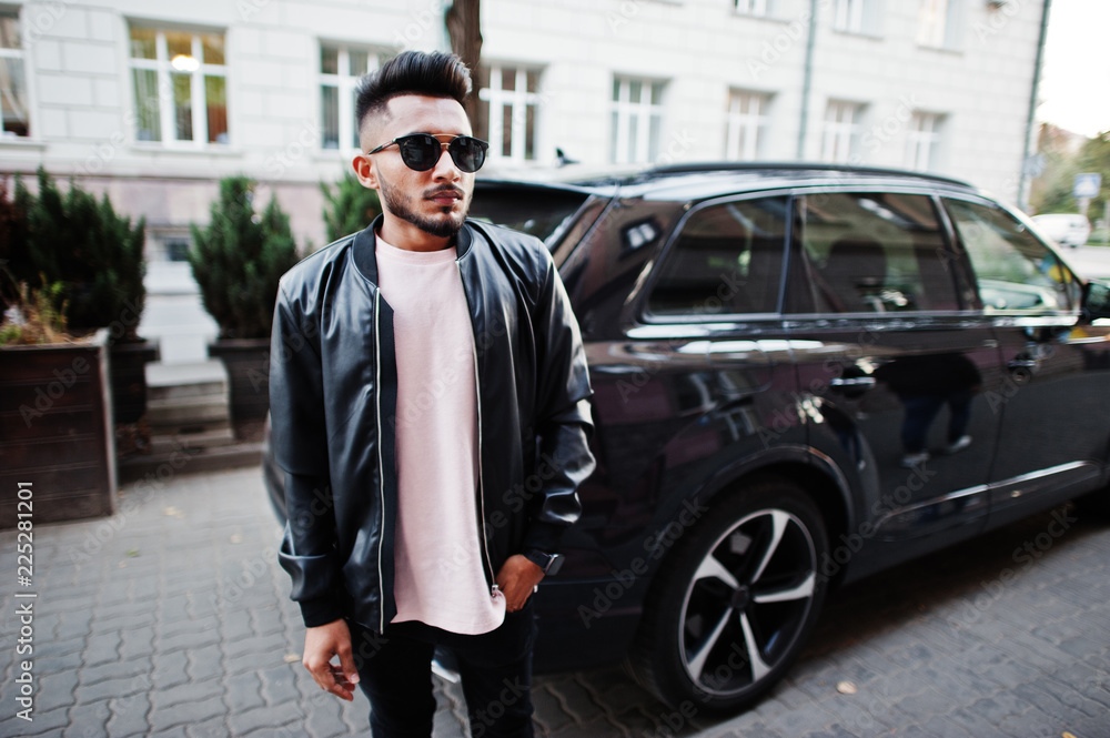 Stylish indian beard man at black leather jacket and sunglasses against business suv car. India model posed outdoor at streets of city.