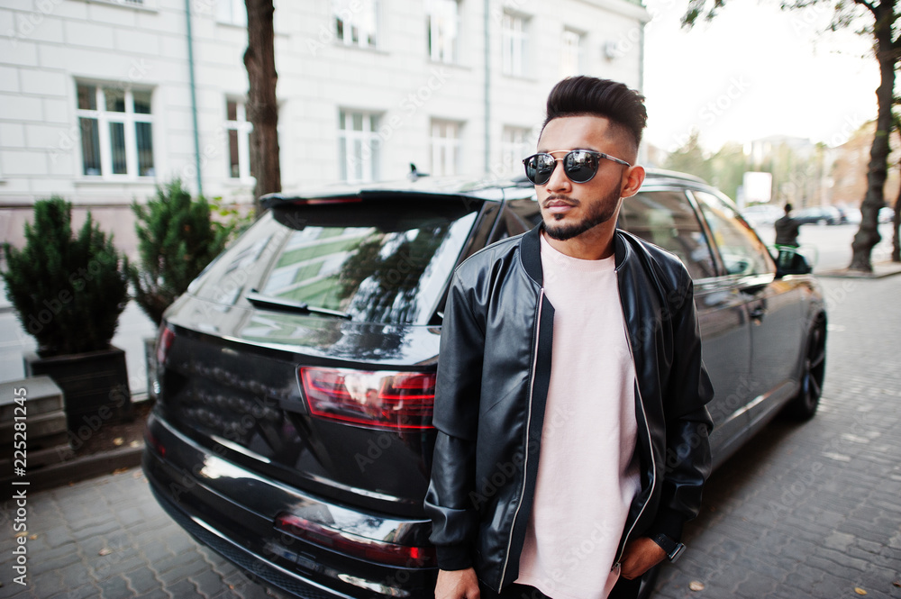 Stylish indian beard man at black leather jacket and sunglasses against business suv car. India model posed outdoor at streets of city.