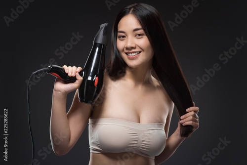 portrait of smiling asian woman with hair dryer looking at camera isolated on black