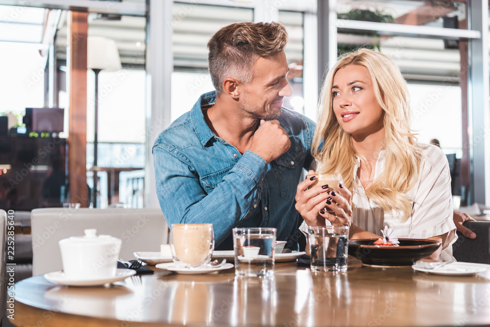 cheerful girlfriend holding cup of coffee and looking at handsome boyfriend at table in cafe cheerful girlfriend holding cup of coffee and looking at handsome boyfriend at table in cafe