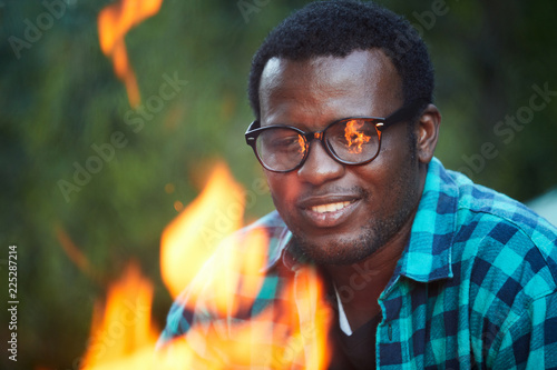 Young man in eyeglases and shirt looking at campfire during backpack trip at leisure photo
