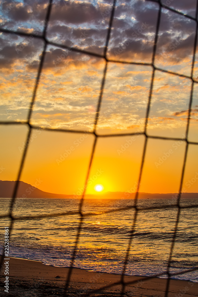 Fototapeta premium View of the sunrise through the volleyball net. Early morning , dramatic sunrise over sea and mountain.