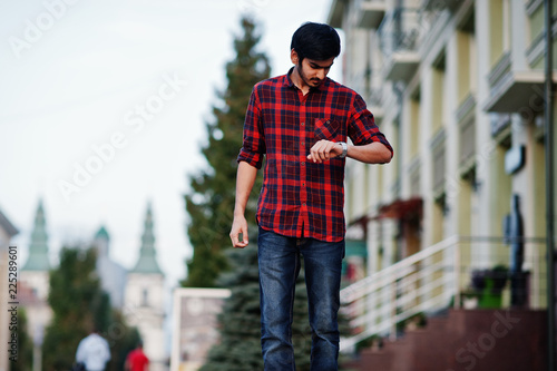 Young indian student man at red checkered shirt and jeans posed at city and looking at his watches. © AS Photo Family