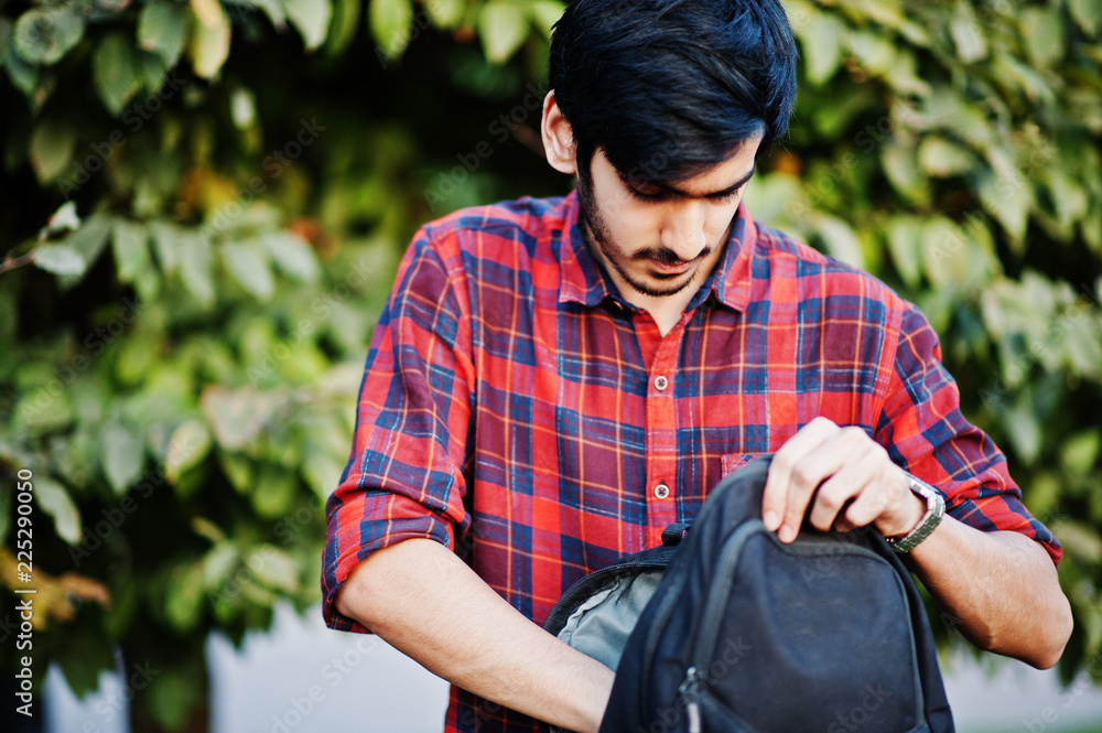 Obraz premium Young indian student man at red checkered shirt and jeans with backpack posed at street.