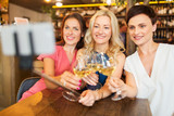 people, technology and lifestyle concept - women drinking wine and taking picture by smartphone on selfie stick  at bar or restaurant
