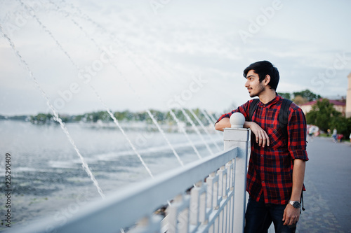 Young indian student man at checkered shirt and jeans with backpack posed on evening city against fountains. © AS Photo Family