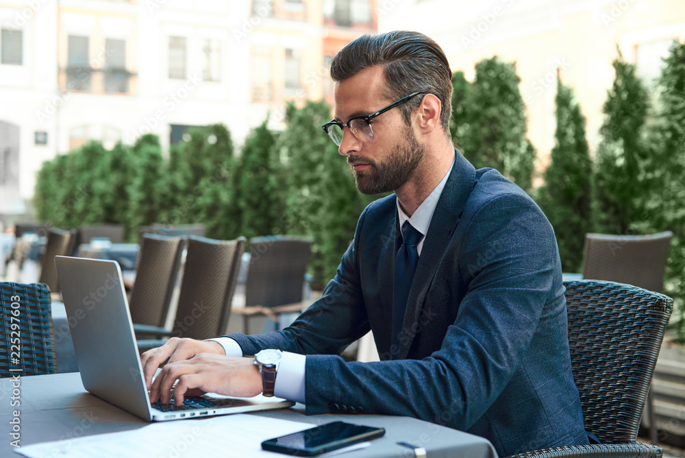 Young businessman with a beard and wearing glasses is sitting and looking at the laptop Young businessman with a beard and wearing glasses is sitting and looking at the laptop