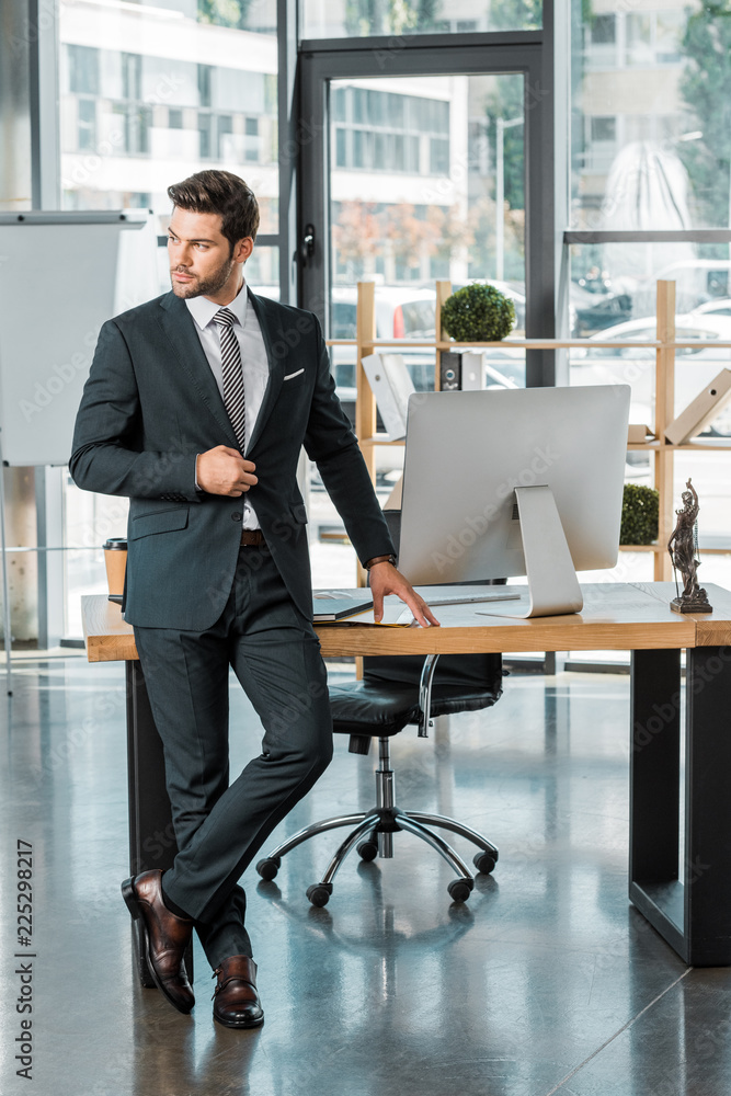 handsome businessman standing near table in office, touching jacket and looking away handsome businessman standing near table in office, touching jacket and looking away