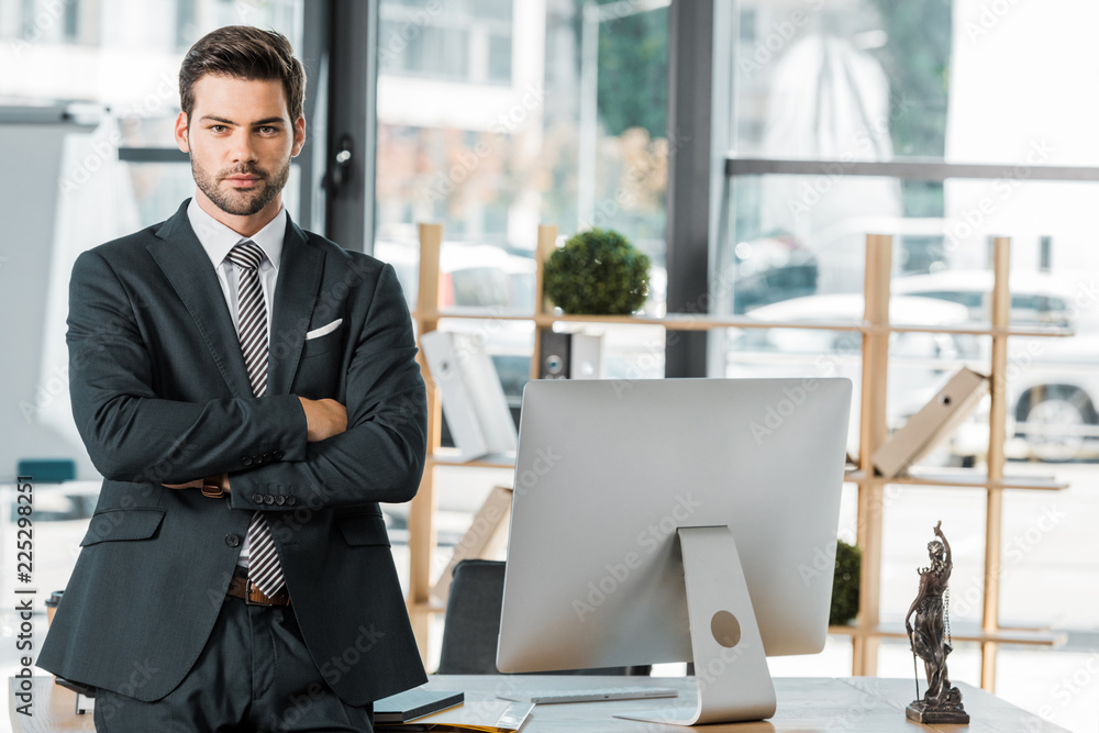 handsome businessman standing with crossed arms near table in office